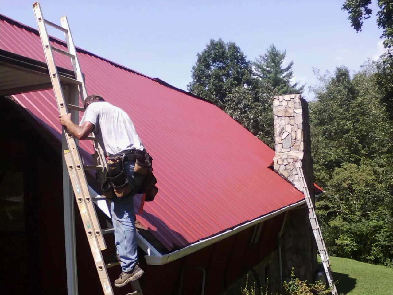 Roofer on a ladder beside a red metal roof and stone chimney — Soffit & Fascia Repair by Roofing Brothers in Poestenkill